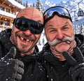 May include: Two individuals wearing black ski jackets, black gloves, and ski goggles. One individual has a mustache and a cigar in their mouth, while the other has missing teeth. Both wear tan-colored balaclavas that cover their heads and necks.