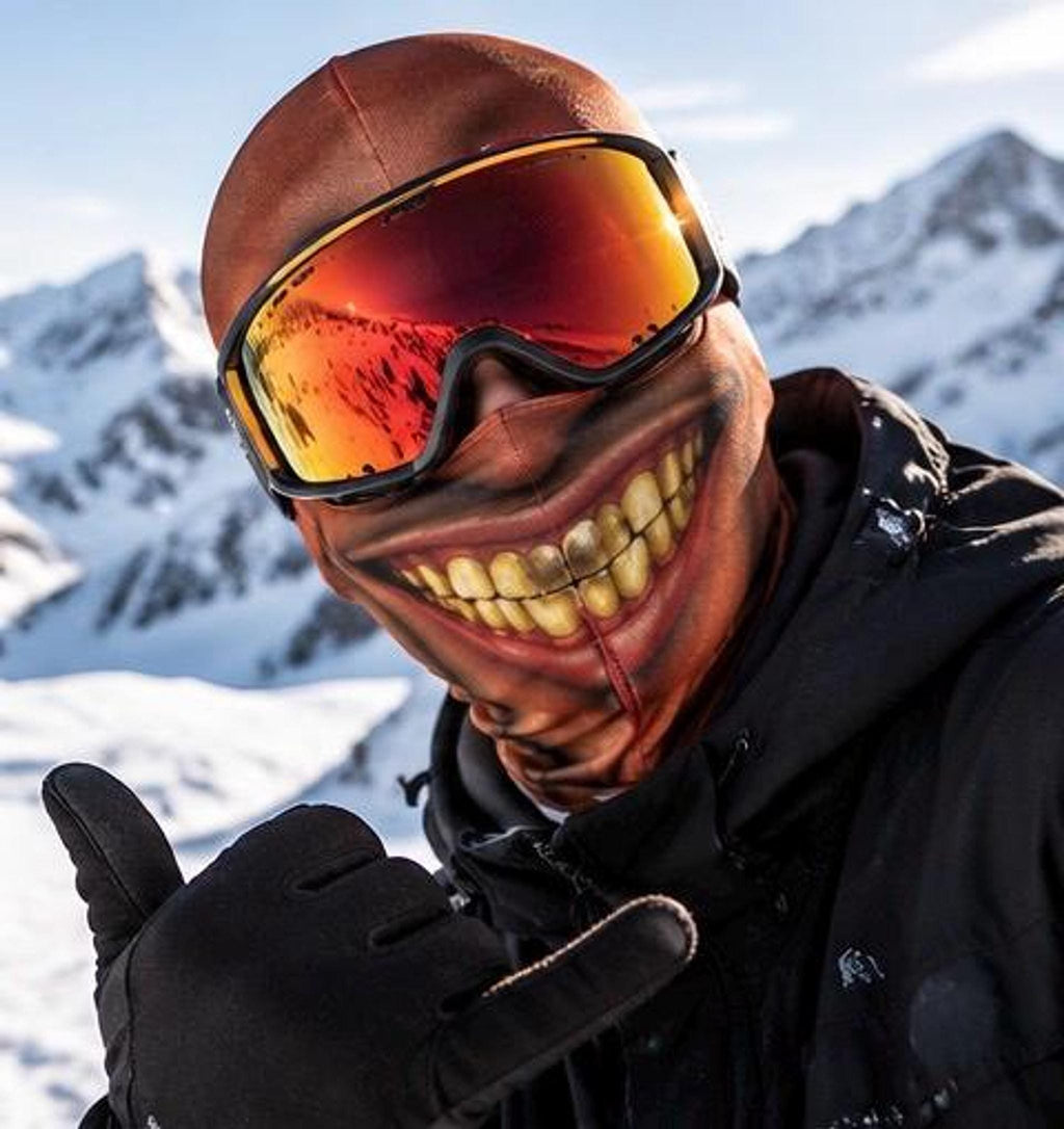 A close-up selfie of a skier on a snowy mountain wearing a brown balaclava printed with a wide, grinning mouth showing yellowed teeth, paired with orange reflective ski goggles.