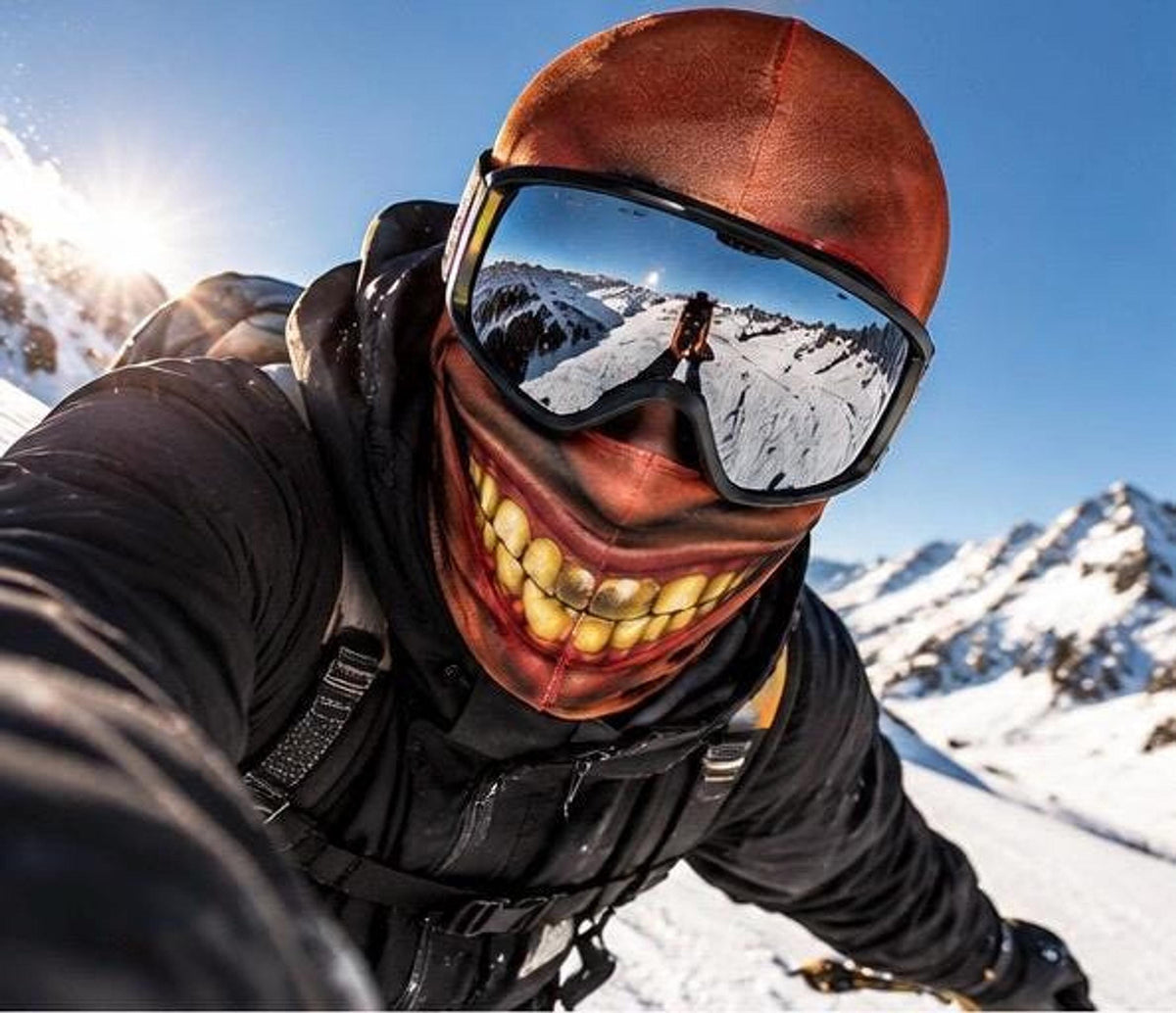 A first-person action shot of a person skiing downhill wearing a reddish-brown balaclava with a graphic of an oversized, toothy grin and mirrored ski goggles reflecting the snowy slope.