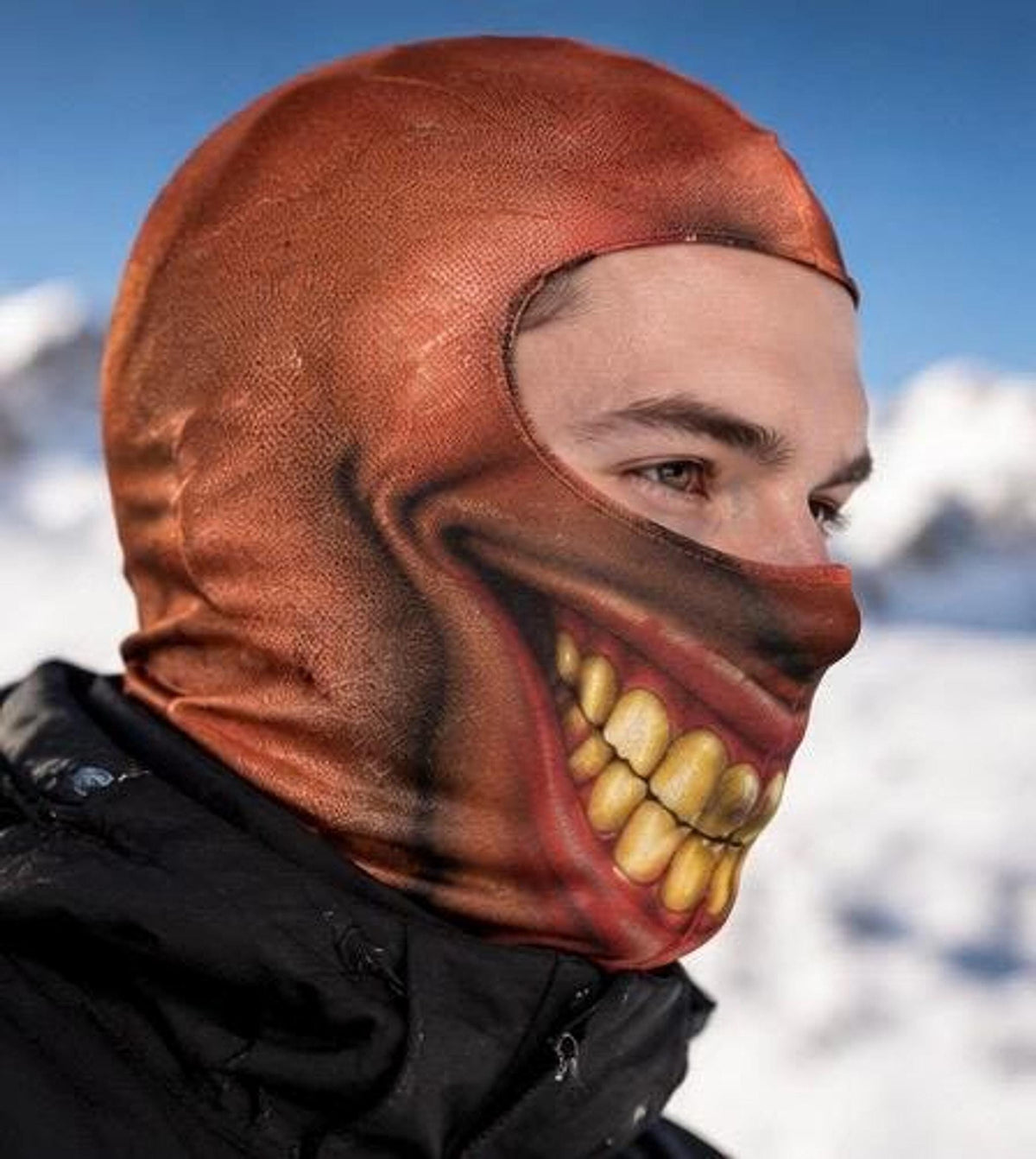 A side profile view of a person wearing the novelty balaclava mask printed with a wide, eerie grin and crooked yellow teeth against an outdoor snowy background.
