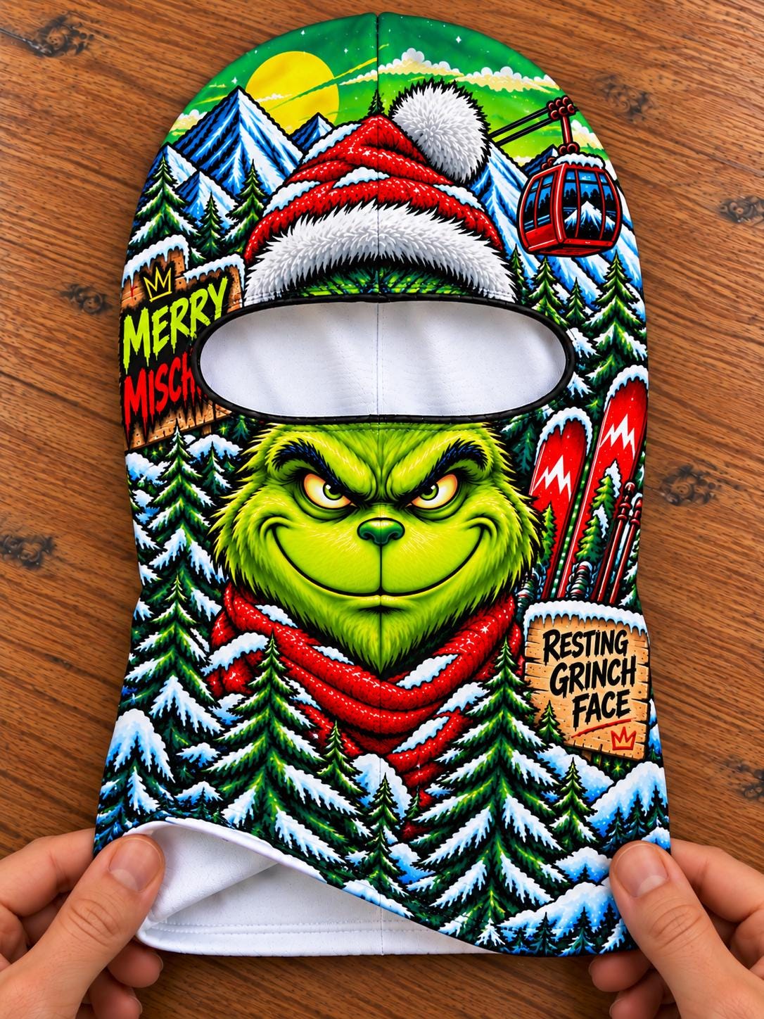 May include: A balaclava featuring a vibrant, full-color graphic of a green character with a mischievous expression, wearing a red scarf. The design includes snowy mountains, pine trees, a red ski lift, and text reading MERRY MISCHIEF and RESTING GRINCH FACE.