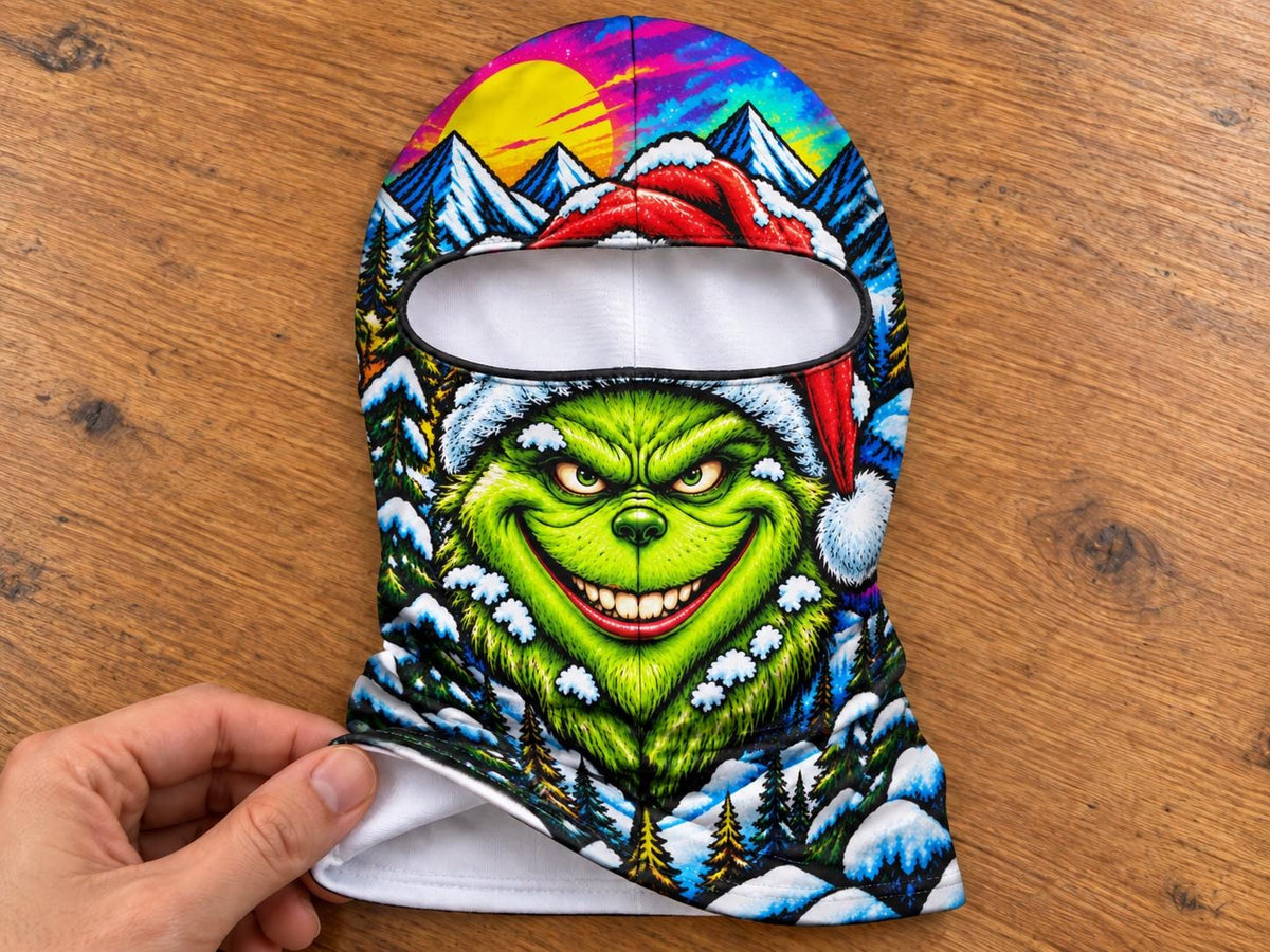 May include: A balaclava featuring a vibrant, colorful graphic print of a green creature with a wide smile wearing a red Santa hat. The background depicts a snowy mountain landscape with pine trees, a bright yellow sun, and a purple sky.
