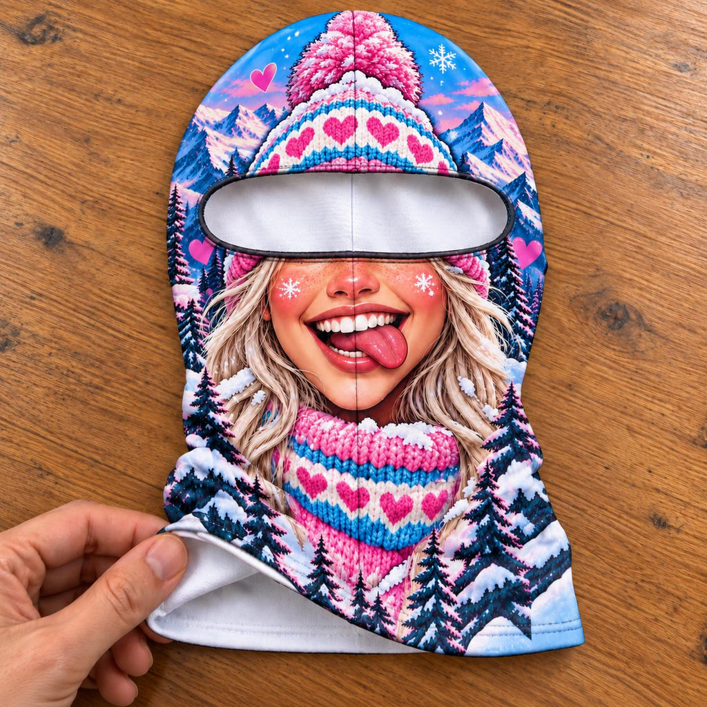 May include: A balaclava featuring a vibrant winter scene with a smiling face, blonde hair, and a pink knit hat with heart patterns. The design includes snowy mountains, dark pine trees, and small pink hearts against a bright blue sky background.