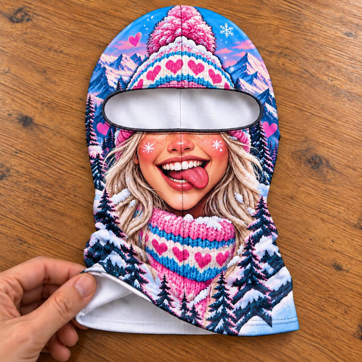 May include: A balaclava featuring a vibrant winter scene with a smiling face, blonde hair, and a pink knit hat with heart patterns. The design includes snowy mountains, dark pine trees, and small pink hearts against a bright blue sky background.