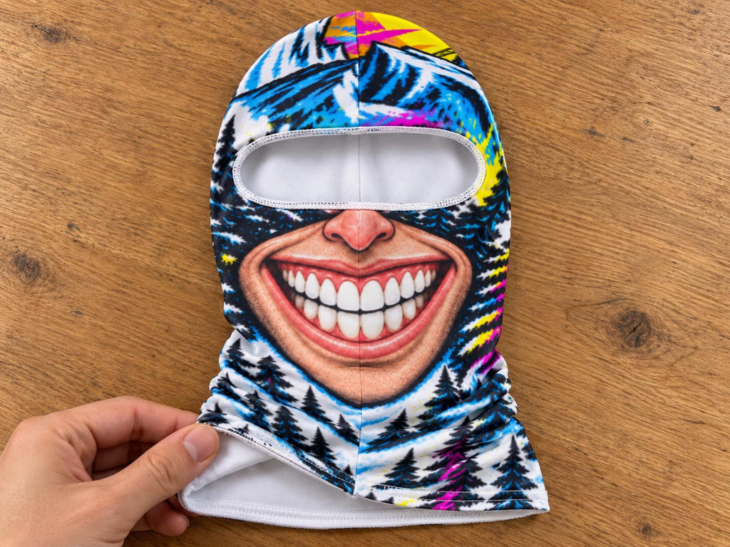 May include: A balaclava featuring a graphic print of a wide, smiling mouth with white teeth, set against a background of blue mountains, dark pine trees, and vibrant yellow and pink accents. The interior lining is solid white fabric.