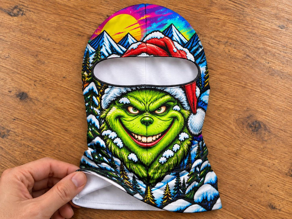 May include: A balaclava featuring a vibrant, colorful graphic print of a green creature with a wide smile wearing a red Santa hat. The background depicts a snowy mountain landscape with pine trees, a bright yellow sun, and a purple sky.