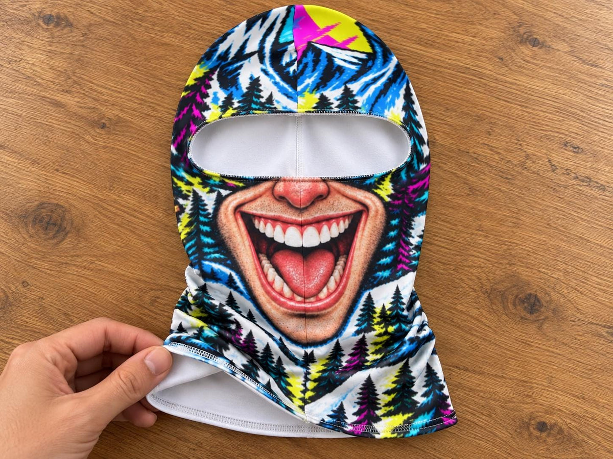 May include: A balaclava featuring a vibrant, multi-colored graphic print of a wide, laughing mouth with visible teeth and tongue, set against a background of stylized pine trees in shades of blue, yellow, pink, and black with a white interior lining.