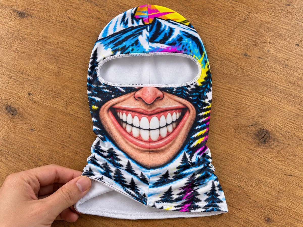 May include: A balaclava featuring a graphic print of a wide, smiling mouth with white teeth, set against a background of blue mountains, dark pine trees, and vibrant yellow and pink accents. The interior lining is solid white fabric.