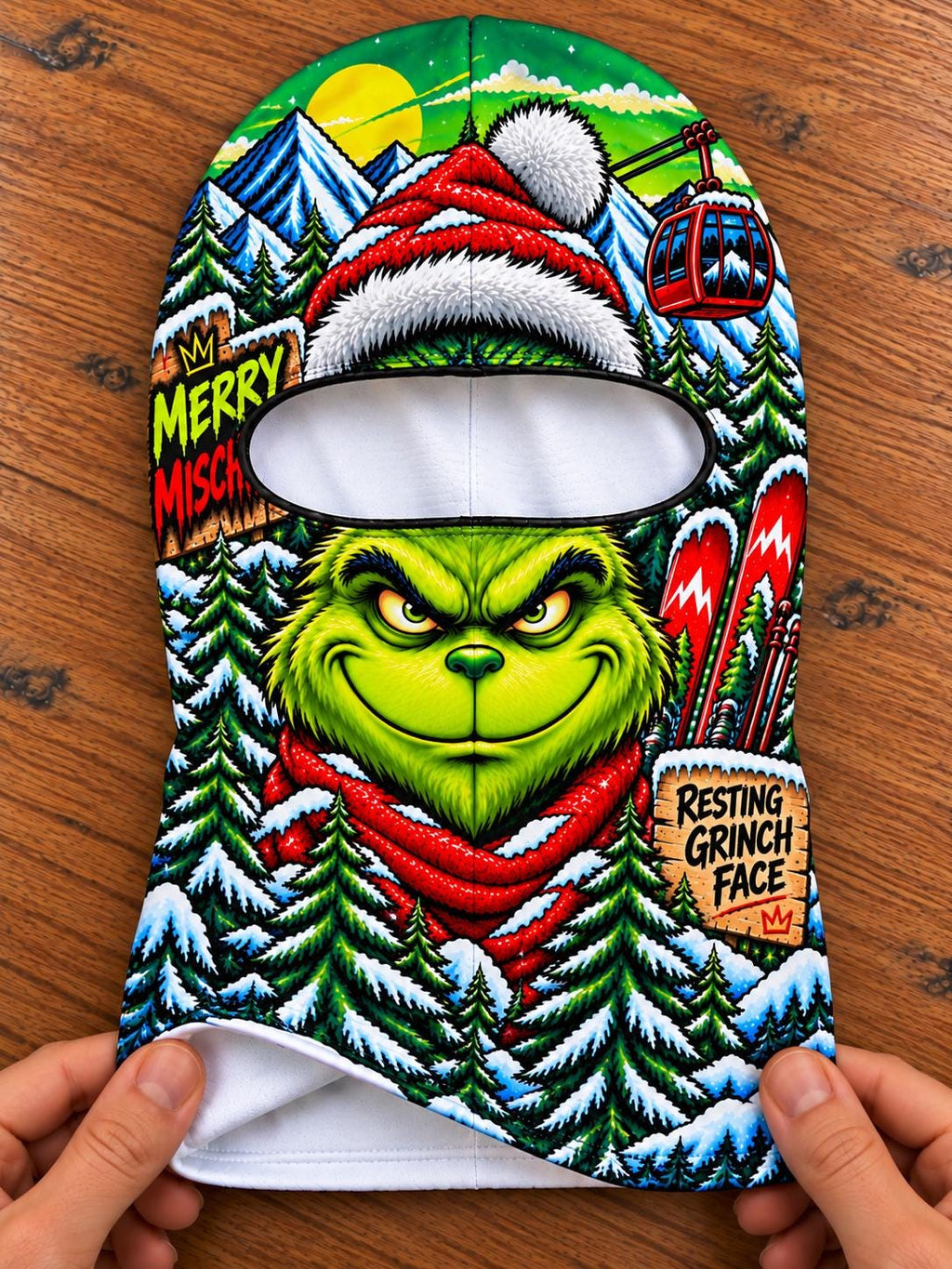 May include: A balaclava featuring a vibrant, full-color graphic of a green character with a mischievous expression, wearing a red scarf. The design includes snowy mountains, pine trees, a red ski lift, and text reading MERRY MISCHIEF and RESTING GRINCH FACE.