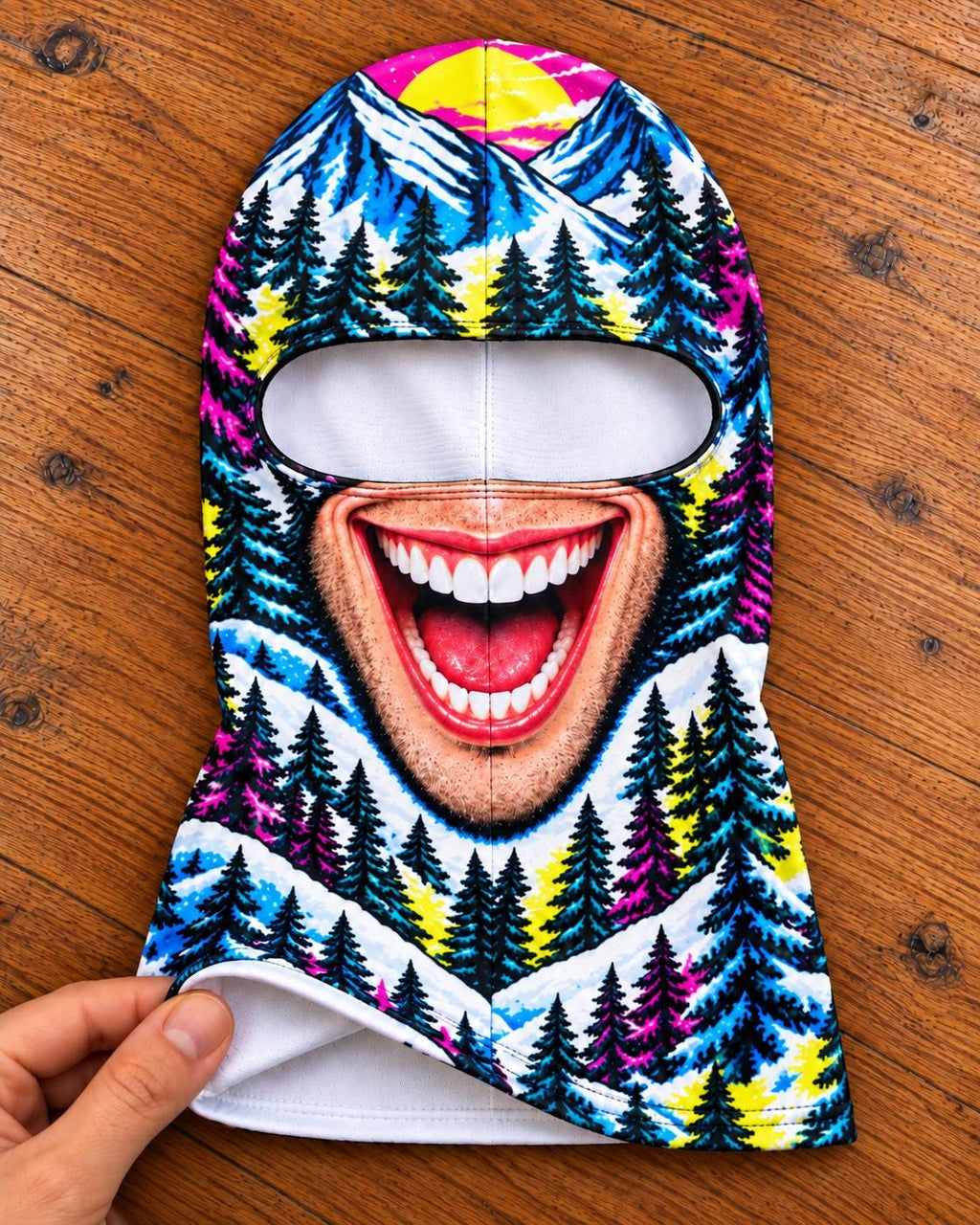 May include: A balaclava featuring a vibrant, colorful graphic print of a snowy mountain landscape with pine trees, a bright yellow sun, and a large, realistic open mouth with white teeth and a red tongue centered below the eye opening.