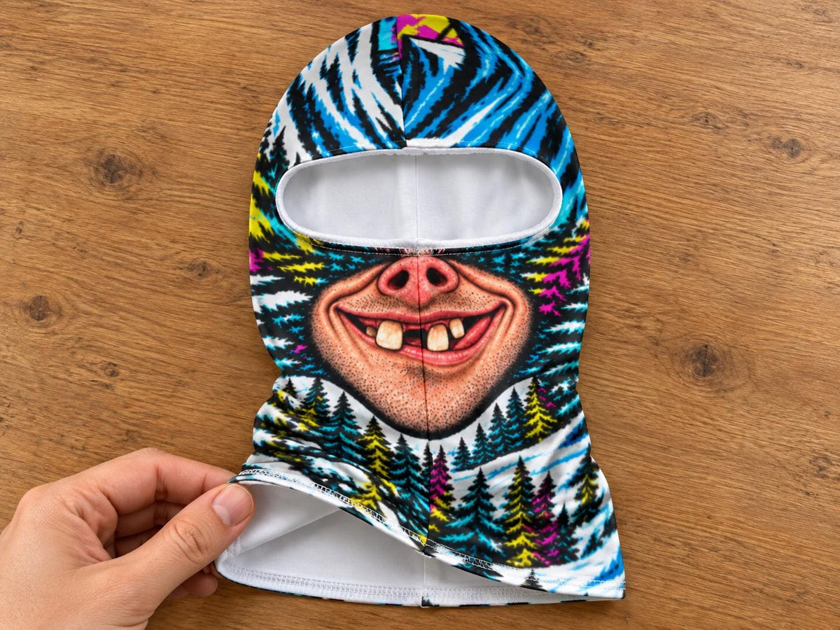 May include: A balaclava featuring a vibrant graphic print of a smiling face with prominent teeth and a pig-like nose, set against a background of stylized blue, white, and yellow pine trees. The interior lining is solid white fabric.