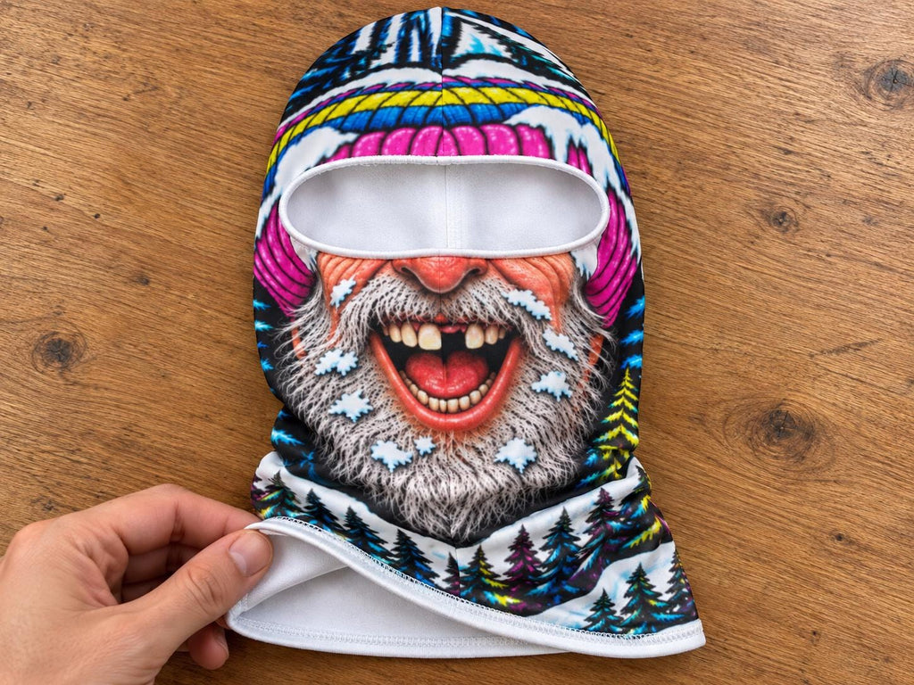 May include: A balaclava featuring a vibrant, full-color graphic print of a laughing, bearded character with a missing tooth. The design includes a colorful winter hat, snowy accents, and a forest of pine trees at the base, all on a white interior.