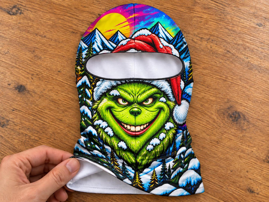 May include: A balaclava featuring a vibrant, colorful graphic print of a green creature with a wide smile wearing a red Santa hat. The background depicts a snowy mountain landscape with pine trees, a bright yellow sun, and a purple sky.