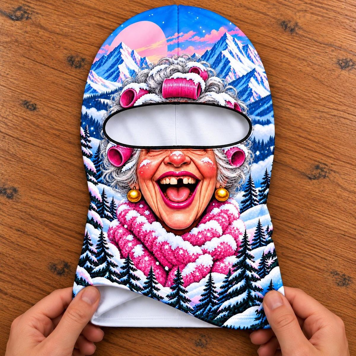May include: A balaclava featuring a colorful graphic print of an older person with gray hair in pink hair rollers, wearing a pink scarf and gold earrings. The background depicts a snowy mountain landscape with pine trees and a pink moon.