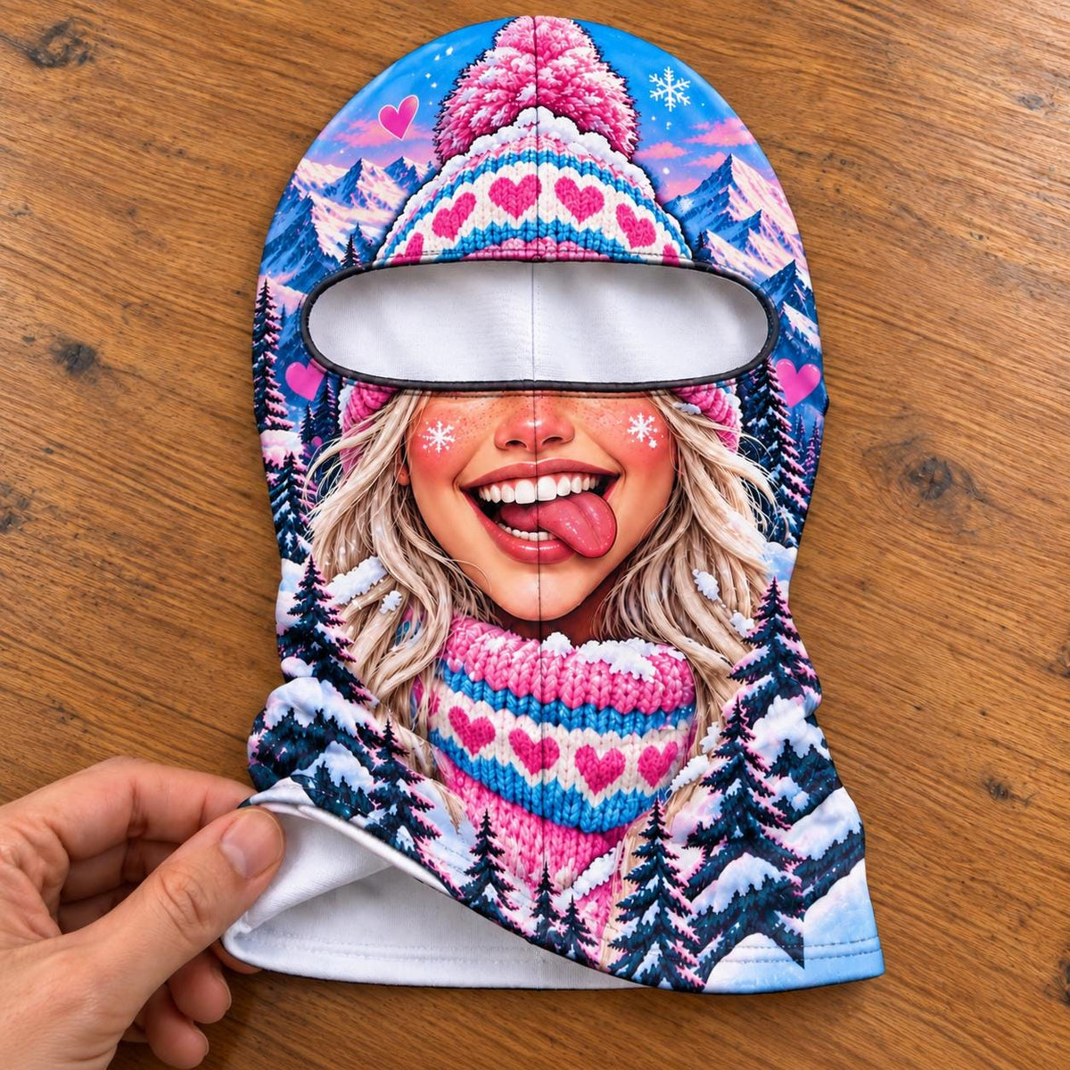 May include: A balaclava featuring a vibrant winter scene with a smiling face, blonde hair, and a pink knit hat with heart patterns. The design includes snowy mountains, dark pine trees, and small pink hearts against a bright blue sky background.