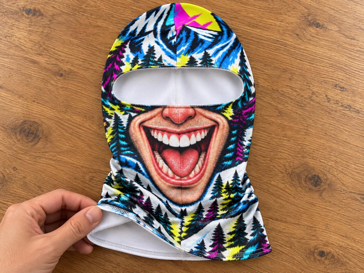 May include: A balaclava featuring a vibrant, multi-colored graphic print of a wide, laughing mouth with visible teeth and tongue, set against a background of stylized pine trees in shades of blue, yellow, pink, and black with a white interior lining.