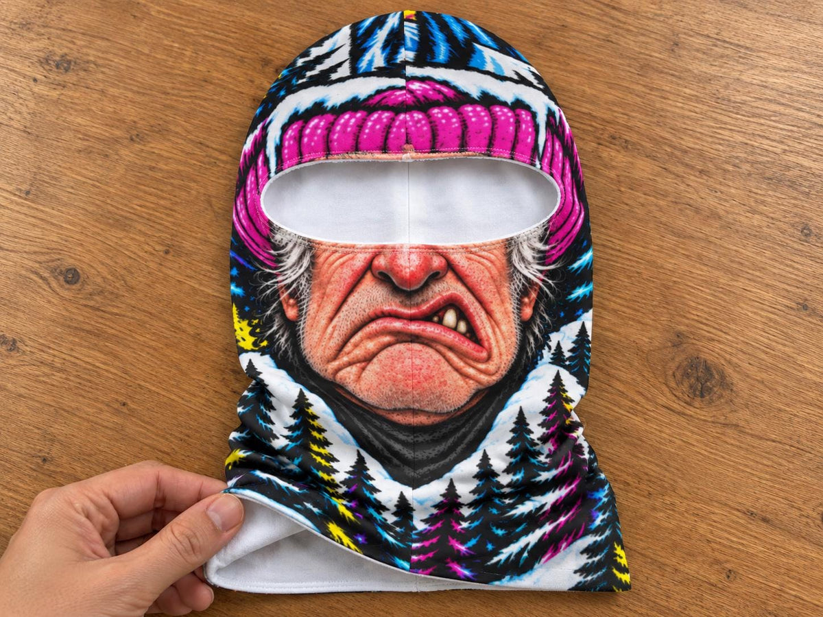 May include: A balaclava featuring a graphic print of a grumpy elderly person wearing a bright pink beanie. The design includes a snowy forest landscape with black pine trees, blue accents, and a white interior lining, creating a vivid, colorful look.