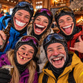 May include: Five individuals wearing black patterned balaclavas with printed realistic facial expressions, including wide grins, missing teeth, and tongues, paired with colorful ski goggles and winter jackets, posing together in a rustic lodge setting.