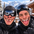 May include: Two individuals wearing black paisley-patterned balaclavas and black ski goggles. One person has a gold tooth and a wide grin, while the other has a gap between their front teeth and their tongue sticking out, both dressed in black jackets.